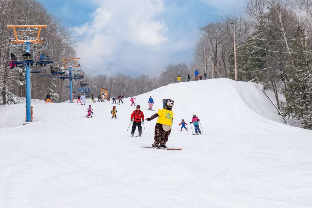 The mascot Rigolo goes down the slope with costumed children. A young snowboarder proudly displays his rechargeable RFID card — Mont Rigaud, ski resort, snowboarding, alpine touring, mountain biking and hiking, family-friendly, in Rigaud, Quebec, near Montreal